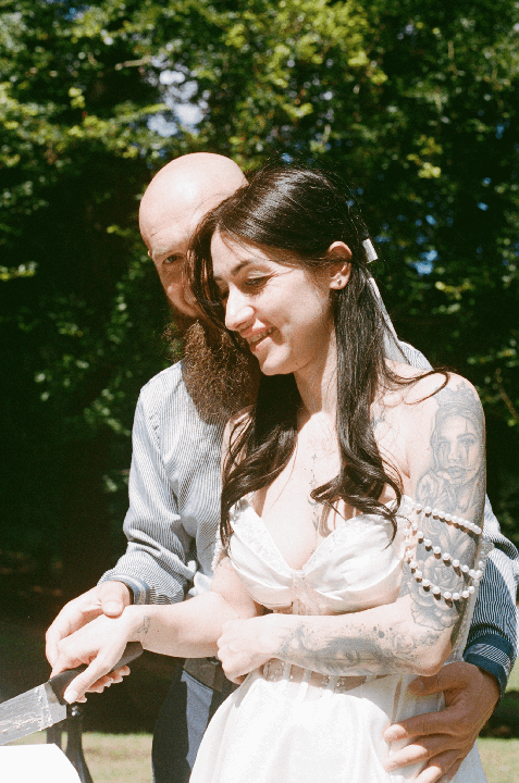 A couple is cutting a cake outdoors, with the woman smiling and wearing a white dress and pearl arm decoration.