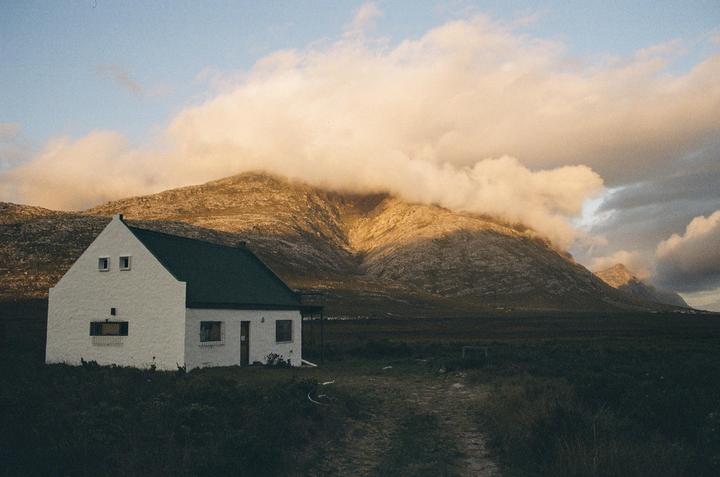 A white house with a dark green roof sits in a field with a large mountain covered in clouds in the background.