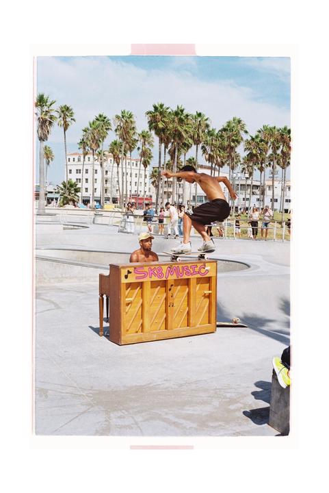 A man on a skateboard jumps over a piano in a skate park with palm trees and buildings in the background.