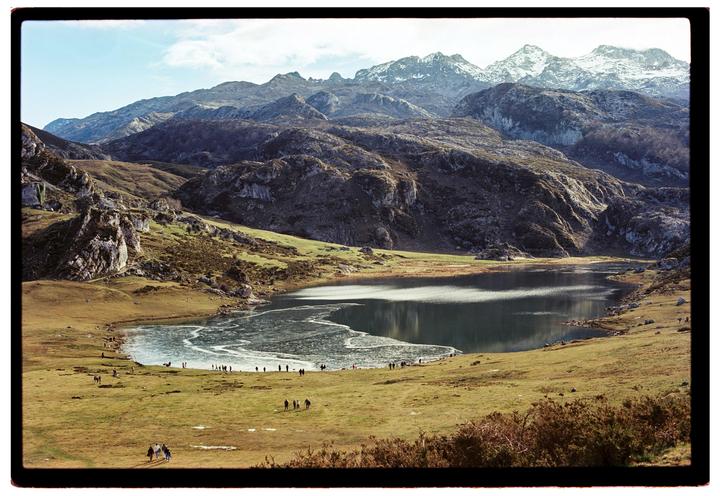 A partially frozen lake is surrounded by grassy hills and snow-capped mountains under a bright sky.
