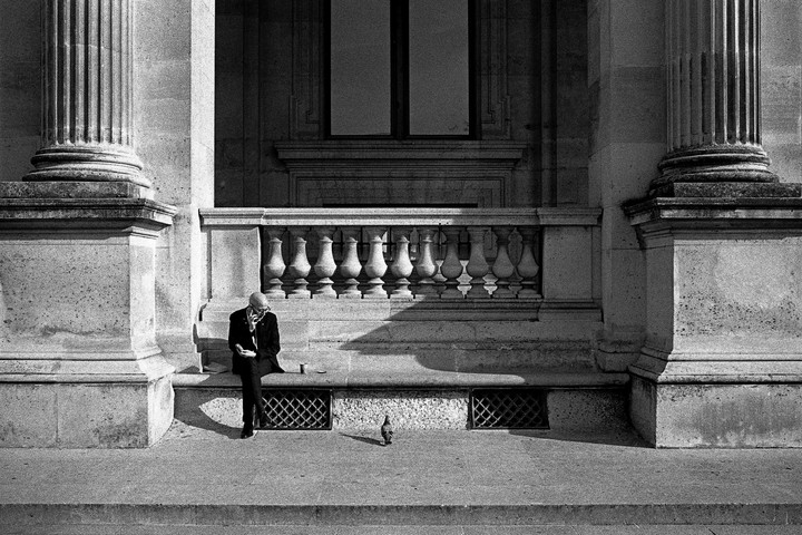 A man sits on a stone ledge next to a pigeon, with large columns and a balustrade behind him.