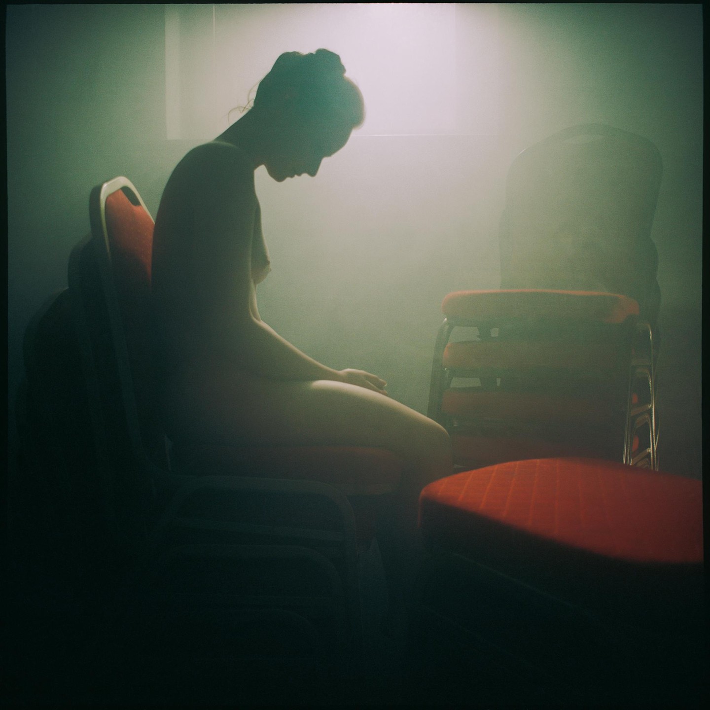 A nude woman sits on a red chair in a hazy room with other chairs.