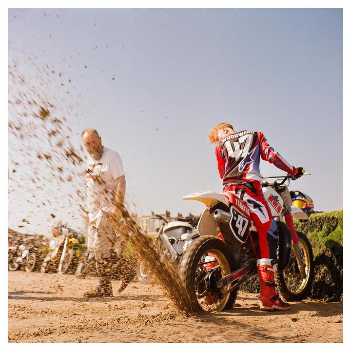 A man on a motorcycle kicks up sand on a beach while another man walks by.