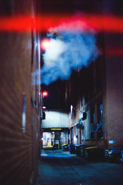 A narrow alleyway at night is illuminated by red and blue light, with smoke rising from a vent.