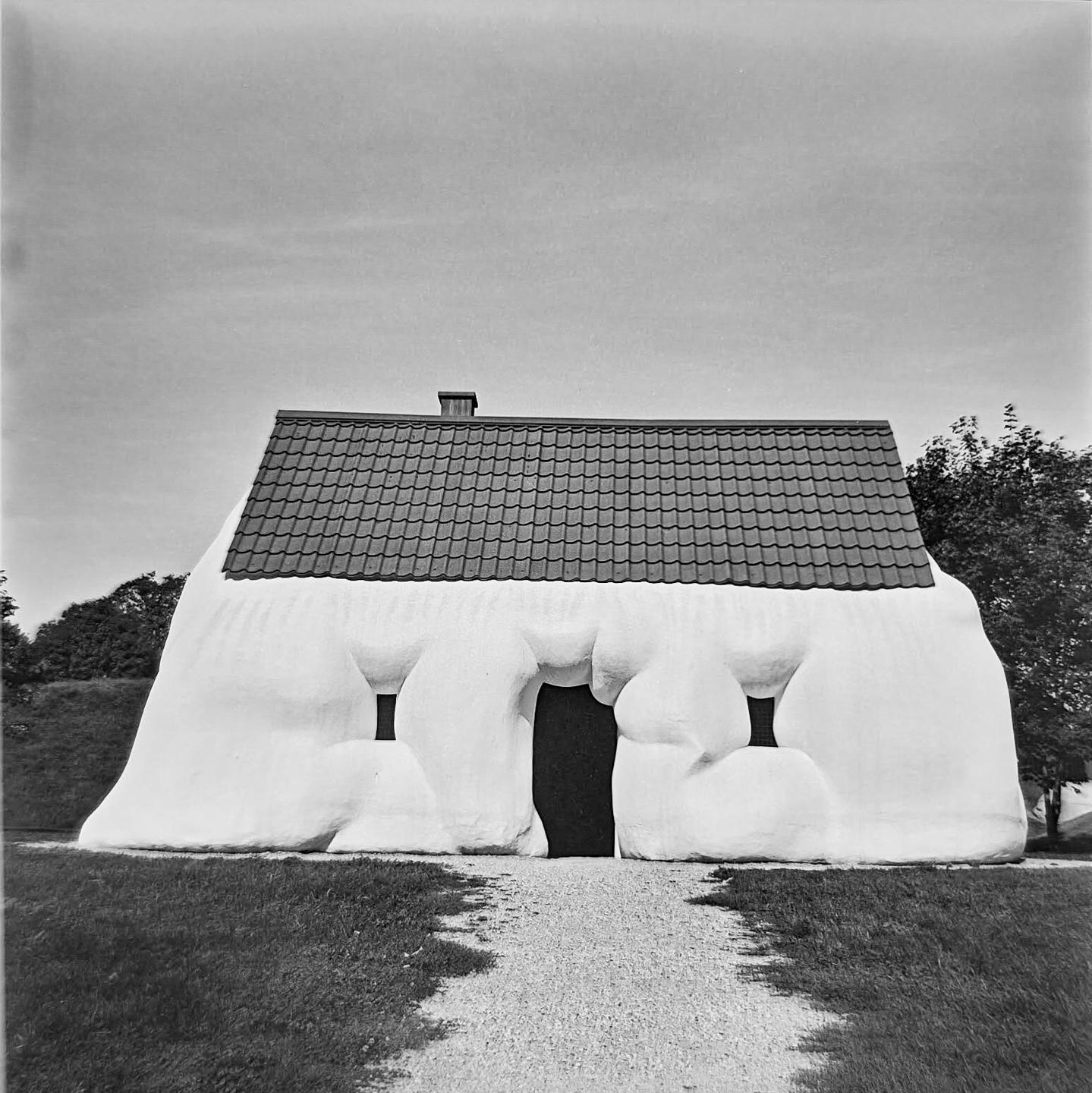 A white house with an unusual, lumpy facade and a tiled roof stands under a clear sky.