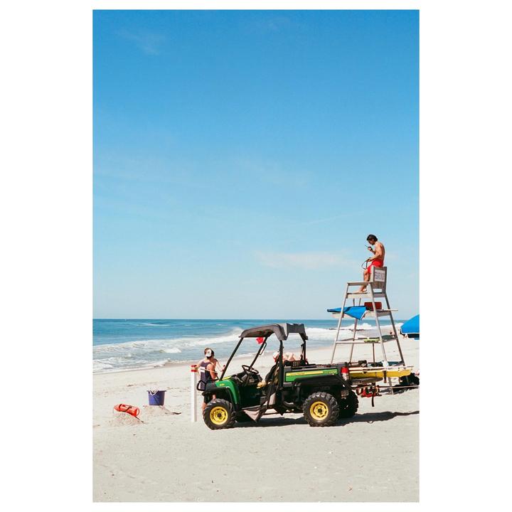 A man sits on a lifeguard chair on a beach next to a utility vehicle and another man.