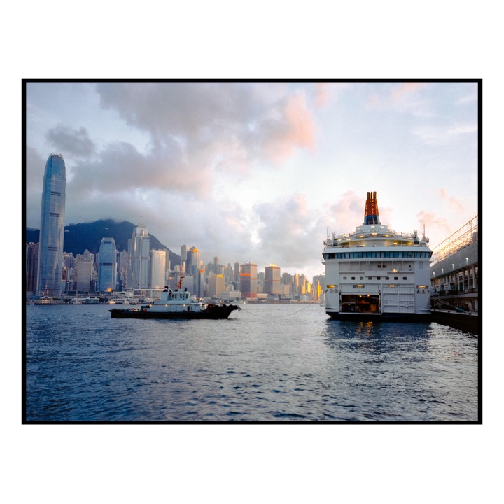 A large white ferry and a smaller tugboat are on the water with a city skyline and mountains in the background under a cloudy sky.