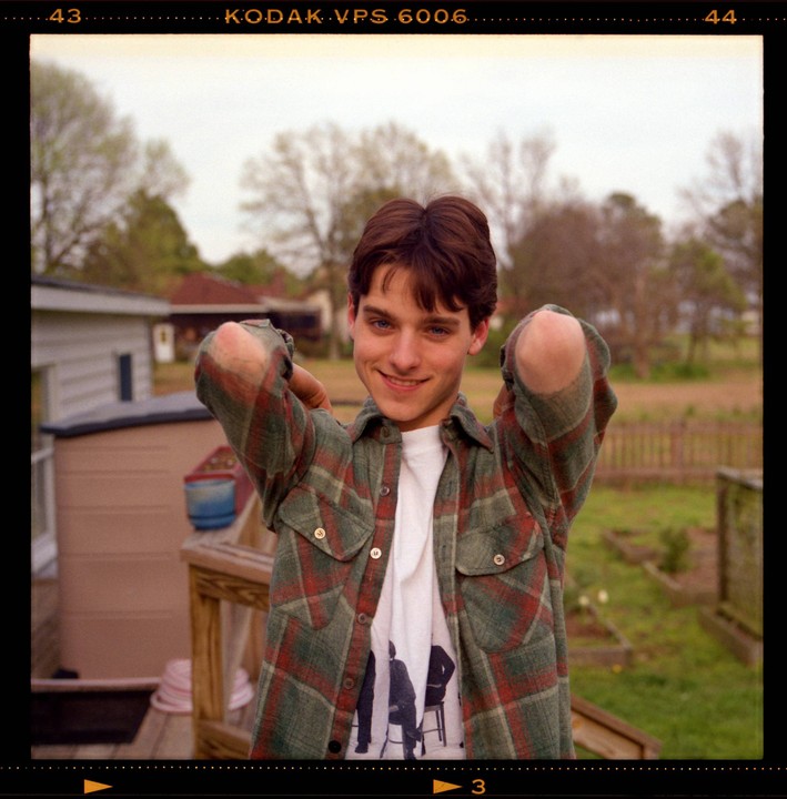 A young man in a plaid shirt smiles at the viewer with his arms raised behind his head.