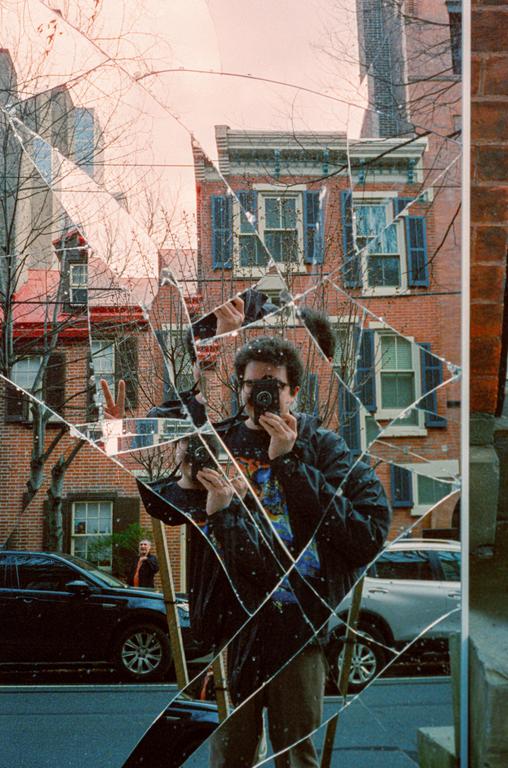 A man takes a self-portrait in a broken mirror on a street with brick buildings and cars.