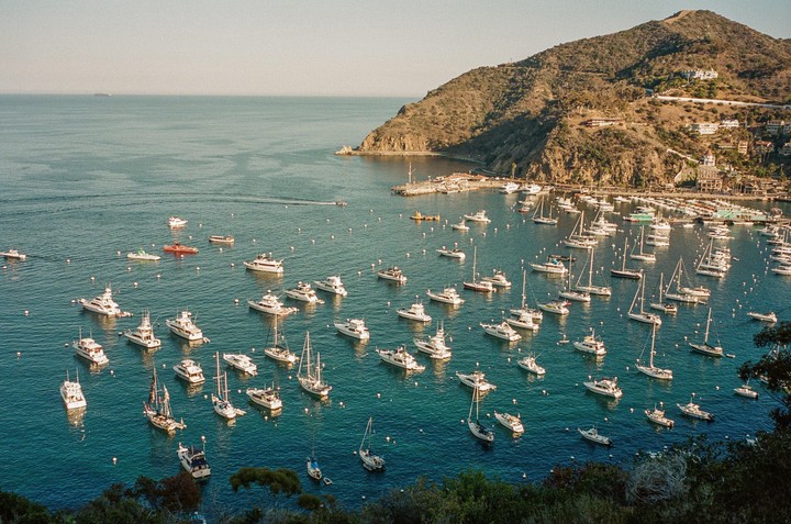 Numerous boats and yachts are moored in a bay next to a mountainous island with buildings on its slopes.