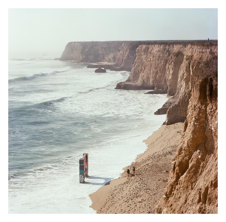 Two people stand on a sandy beach next to a cliff with waves crashing on the shore.