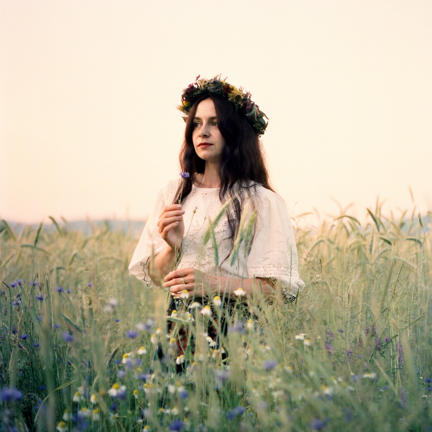 A woman wearing a flower crown stands in a field of wildflowers and wheat.