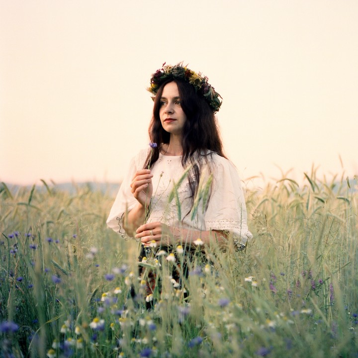 A woman wearing a flower crown stands in a field of wildflowers and wheat.