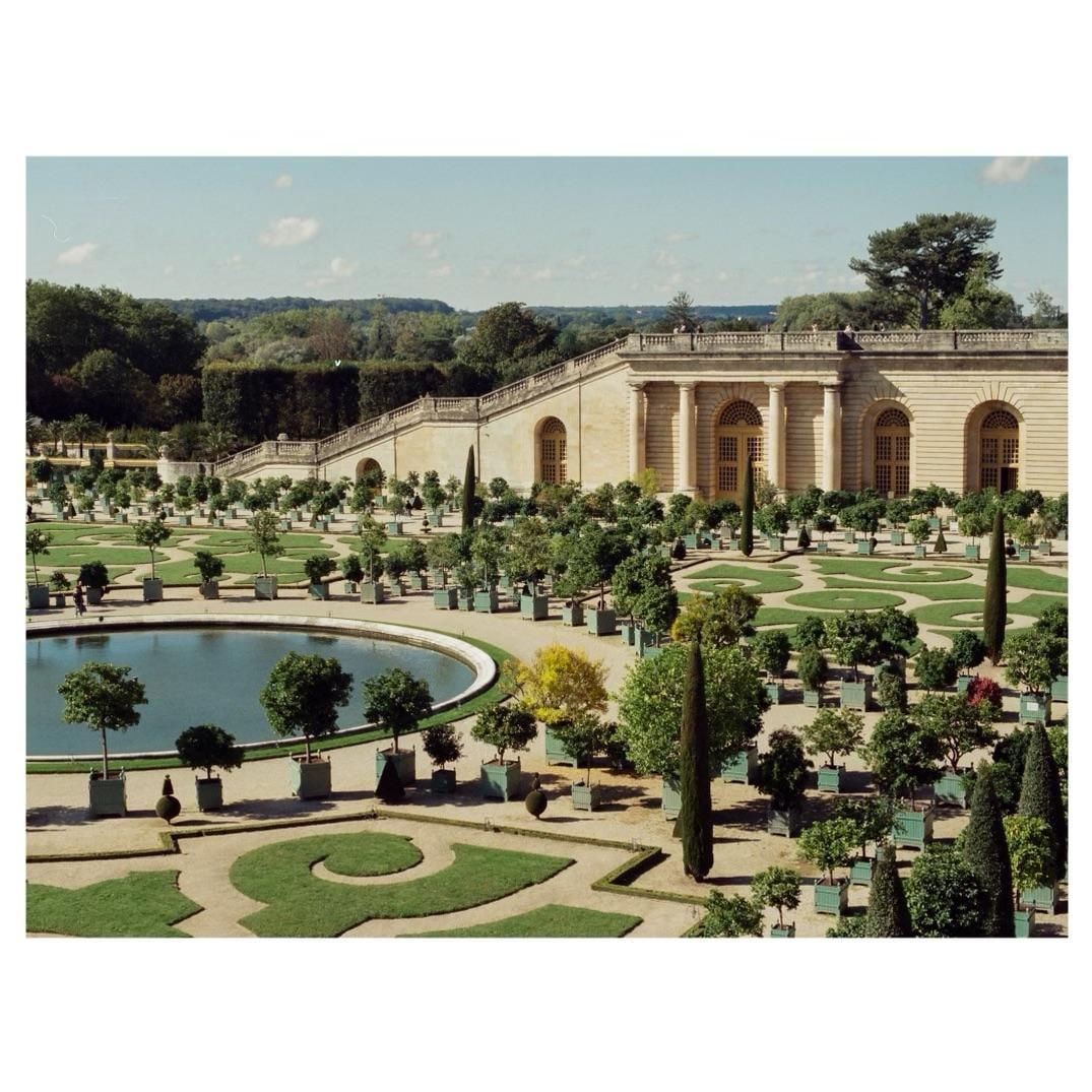 A large formal garden with a circular pond and a grand building with columns and arched windows in the background.