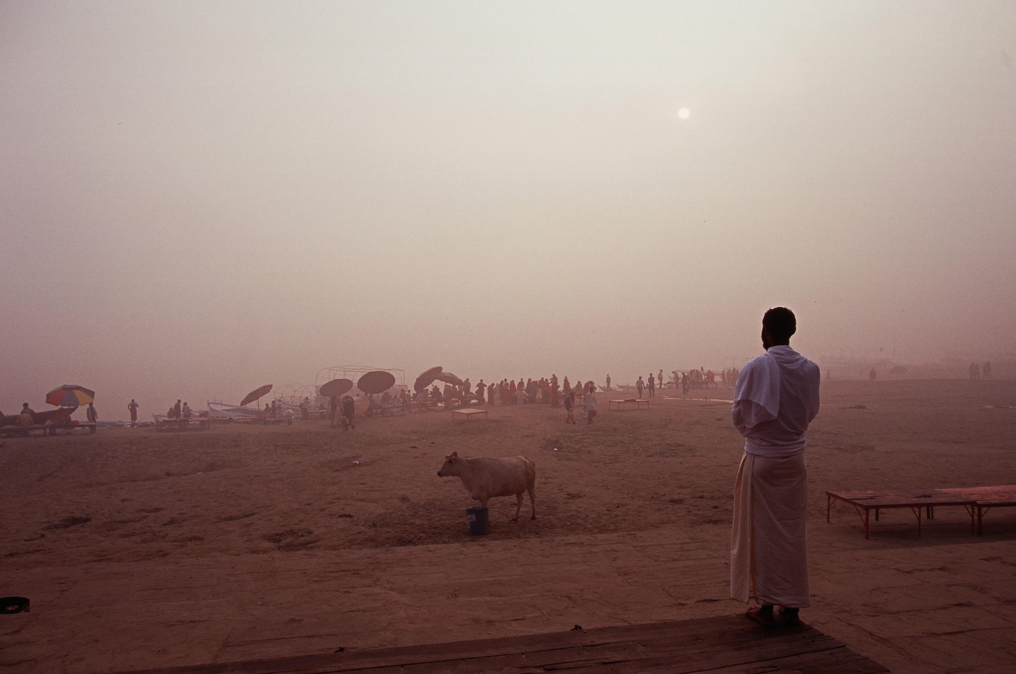 A man in traditional clothing stands on a sandy bank looking out at a hazy river with many people and a cow.
