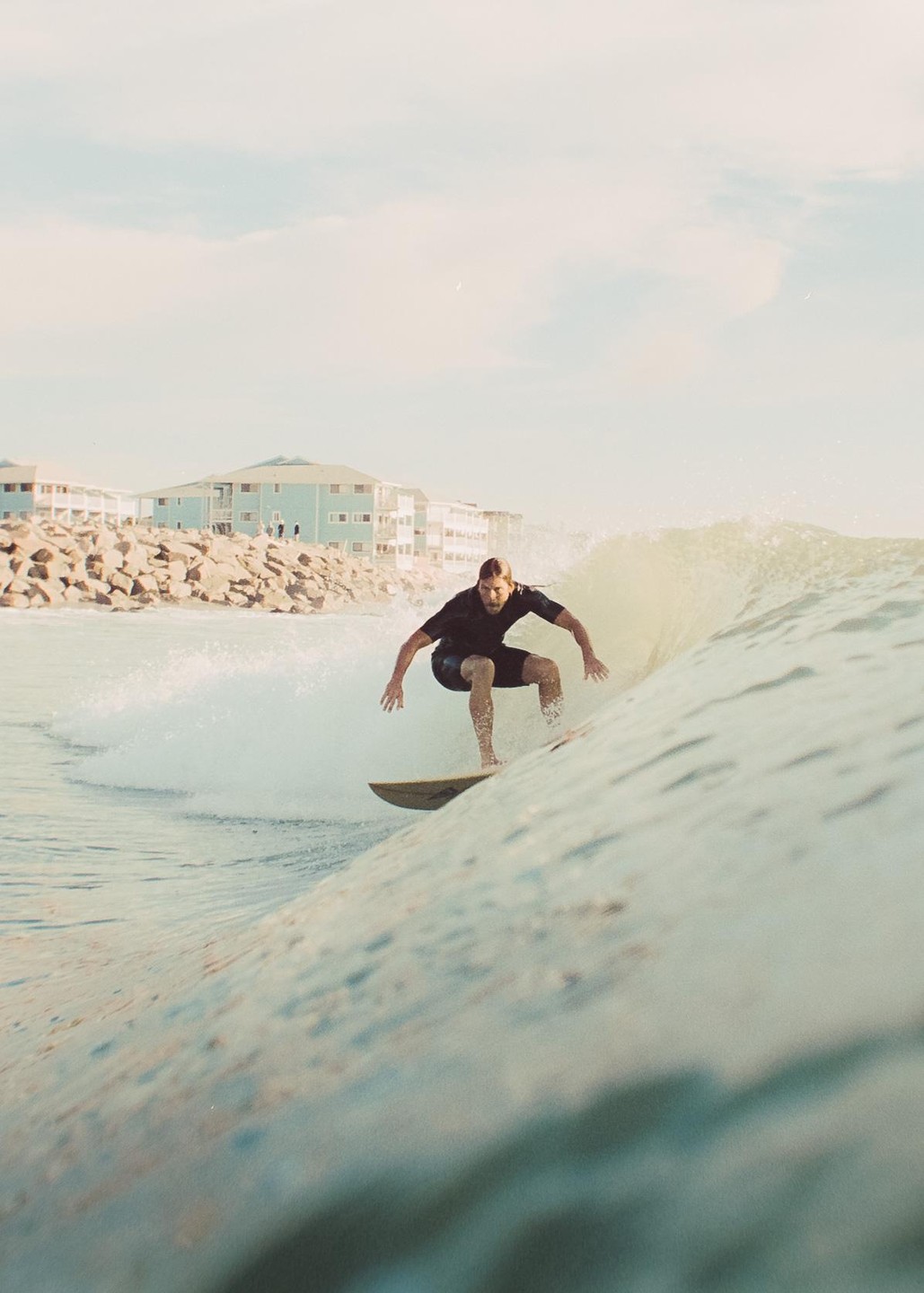 A man on a surfboard rides a wave with buildings and rocks in the background.
