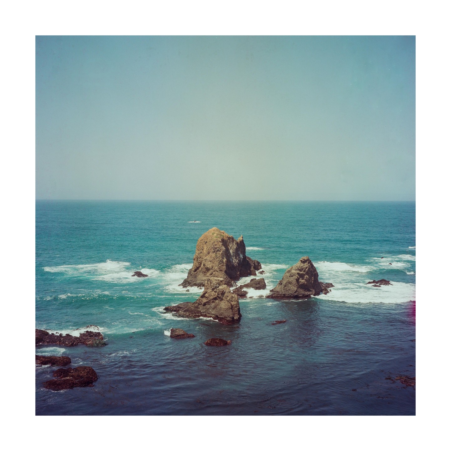 Large rock formations rise from the ocean with waves crashing around them under a hazy sky.
