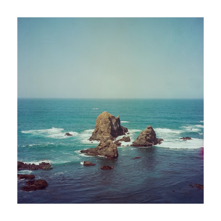 Large rock formations rise from the ocean with waves crashing around them under a hazy sky.