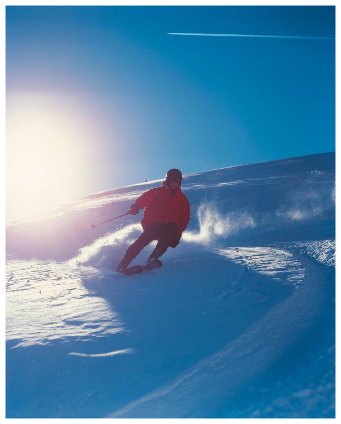 A skier in a red jacket carves a turn on a snowy mountain slope under a bright sun and blue sky.