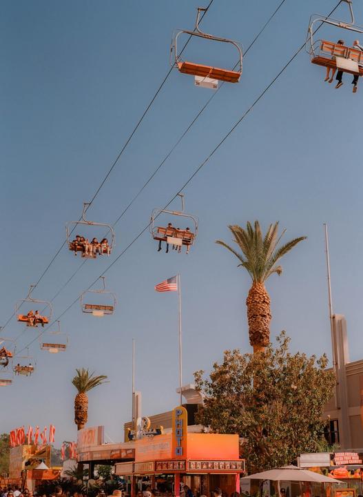 People ride a ski lift over a fairground with palm trees and an American flag.
