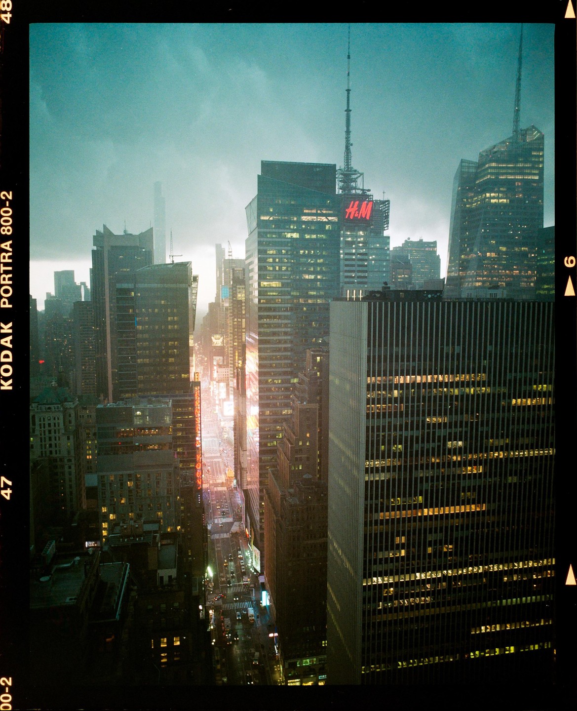 A high-angle view shows a street lined with skyscrapers and illuminated billboards under a cloudy sky.