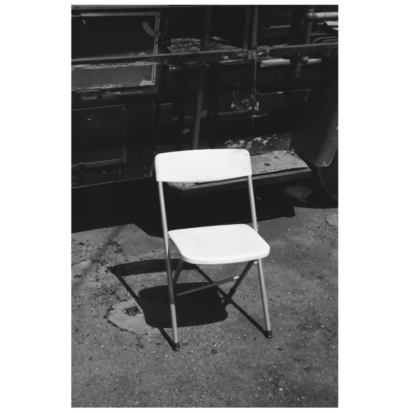 A white folding chair sits on a concrete surface in front of a dark, weathered vehicle.
