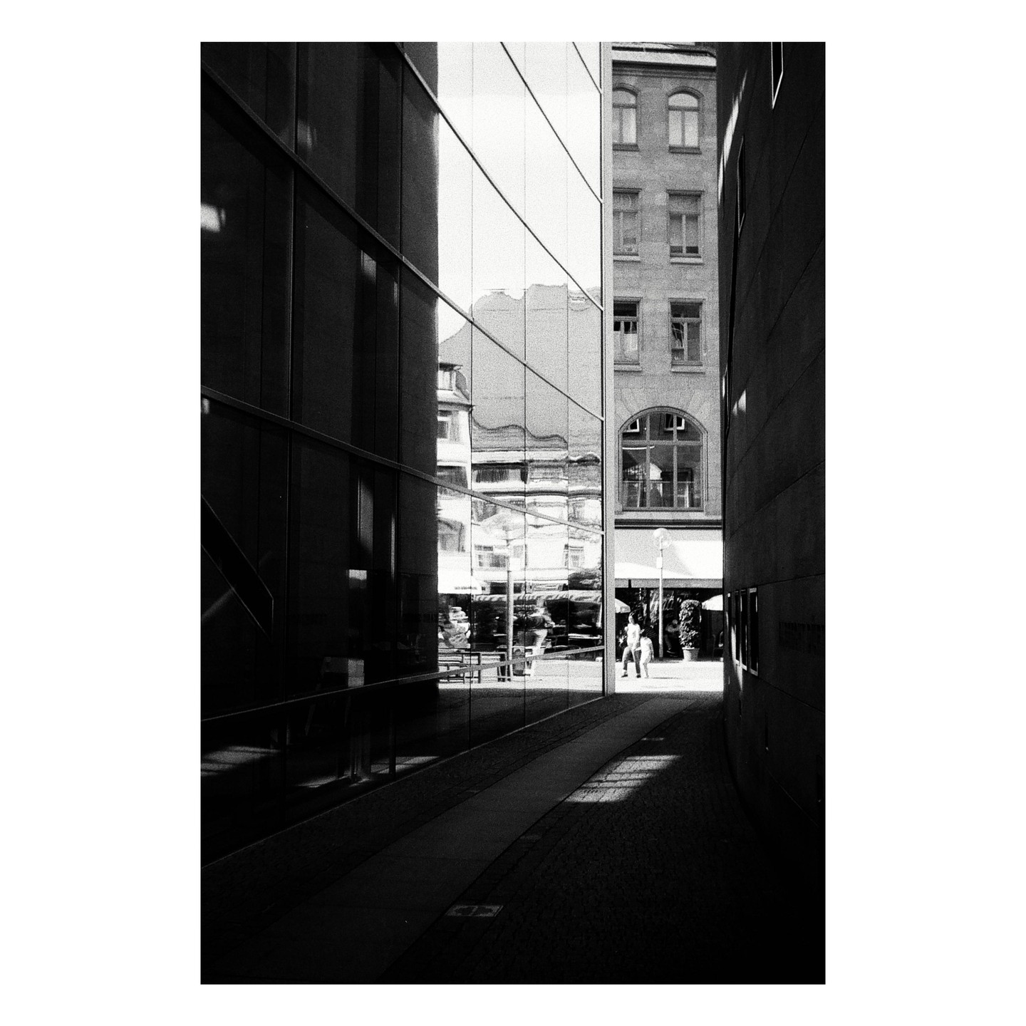 A narrow alleyway is flanked by a modern building with reflective windows and an older building with many windows, with two people walking in the distance.