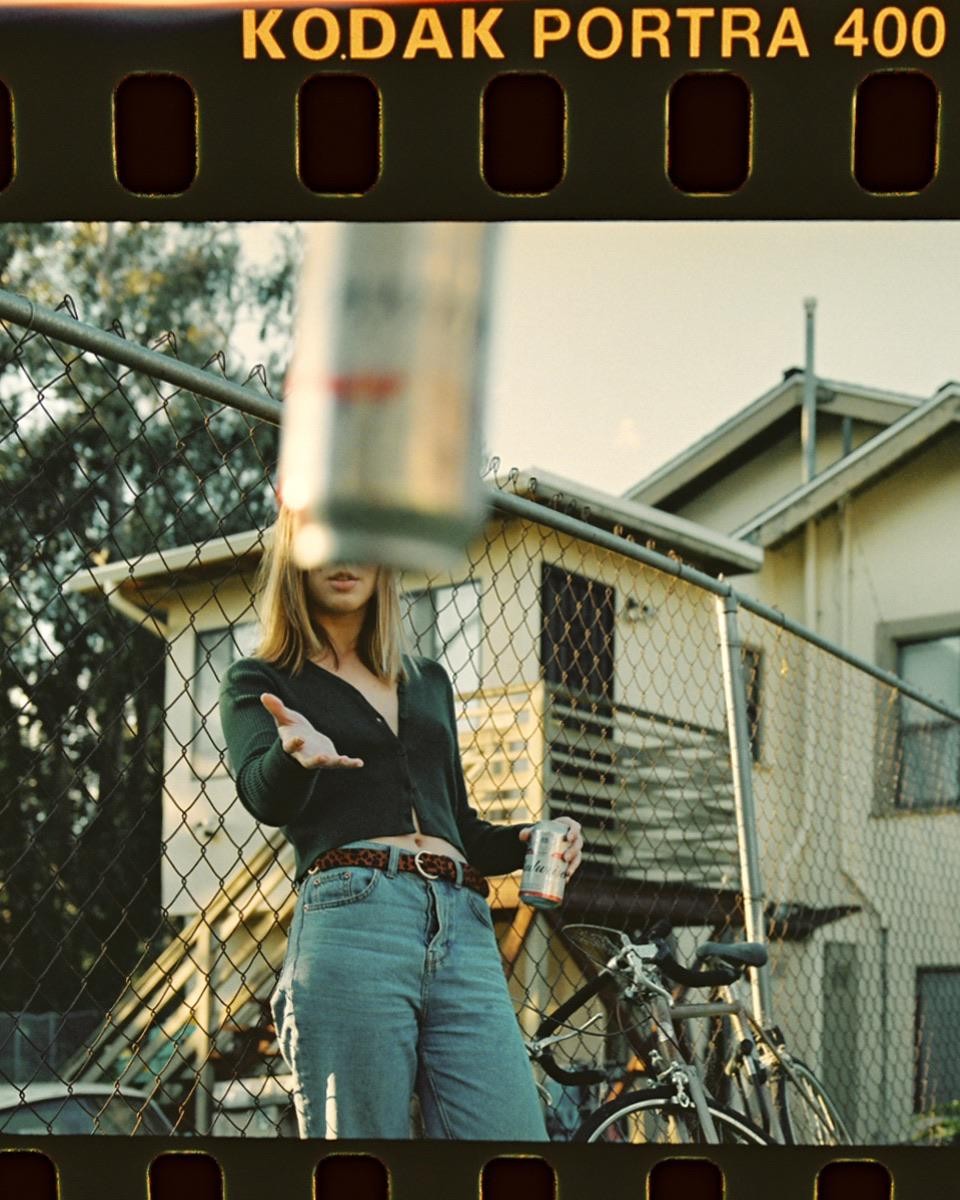 A woman stands in front of a chain link fence, holding a can and reaching out her hand, with a bicycle and a house in the background.