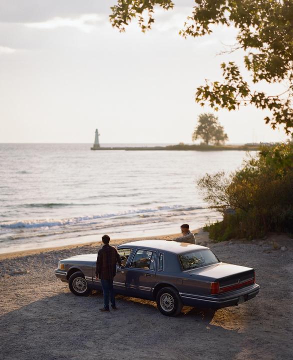 Two men stand next to a car parked on a beach with a lighthouse in the distance.