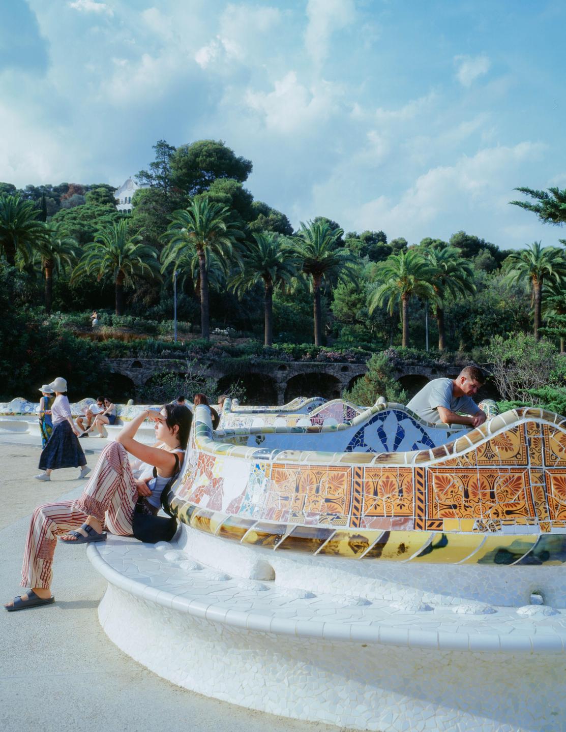 A woman sits on a mosaic bench in a park with palm trees and a man leaning over the bench in the background.
