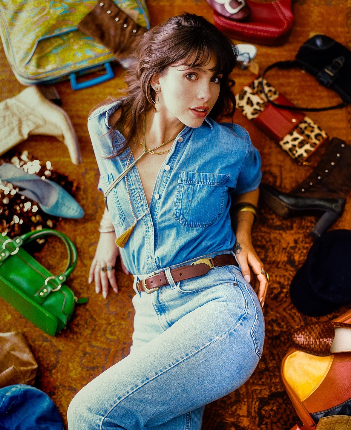 A woman in a denim shirt and jeans is lying on a patterned rug surrounded by various accessories.