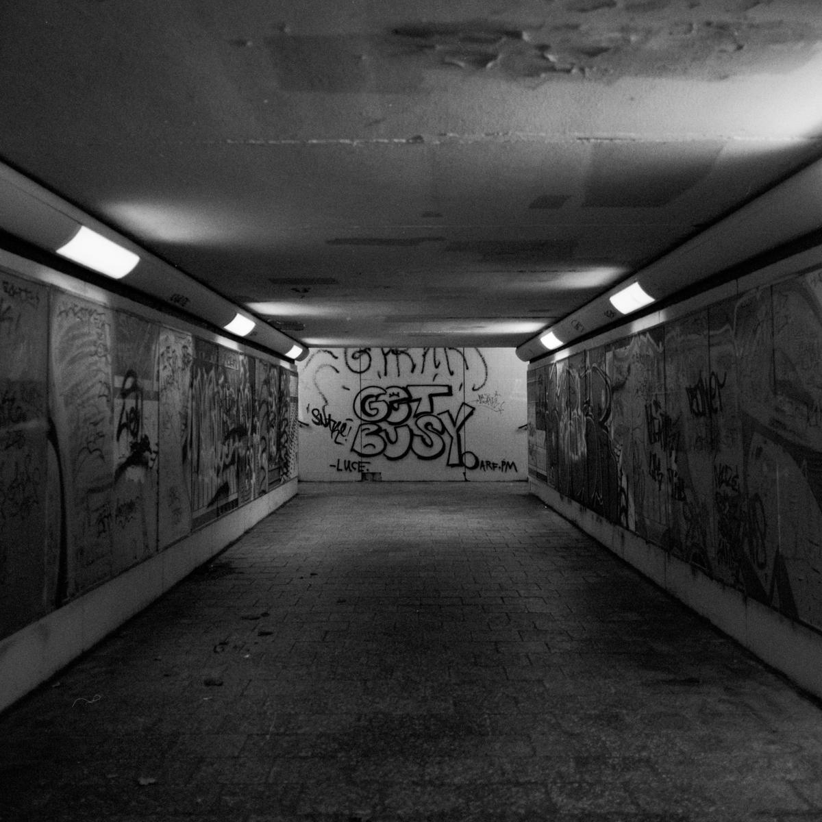 A graffiti-covered pedestrian tunnel is illuminated by fluorescent lights.