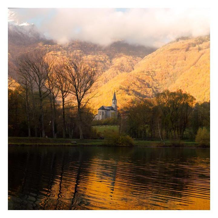 A church with a tall steeple sits on a hill overlooking a lake, surrounded by trees and mountains with autumn foliage.