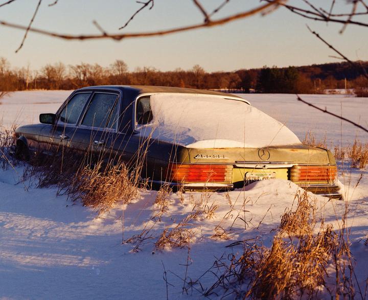 A snow-covered car is parked in a field with dry grass and trees in the background.