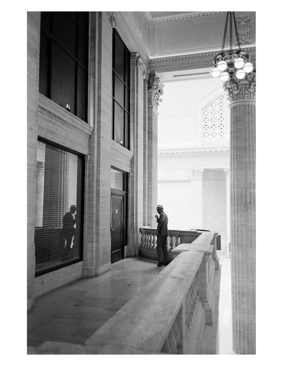 A man in a suit and hat stands on a balcony inside a grand building with tall columns and large windows.