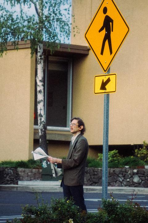 A man in a suit and glasses stands on a street corner holding a newspaper next to a pedestrian crossing sign.