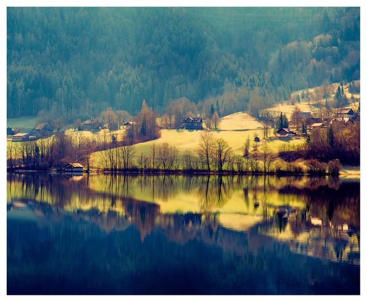 A village with houses and trees is reflected in a lake with a forest in the background.