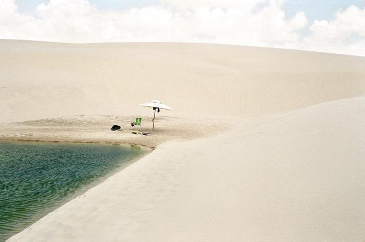 A white umbrella and a green chair sit on a sandy bank next to a body of water, surrounded by sand dunes under a cloudy sky.