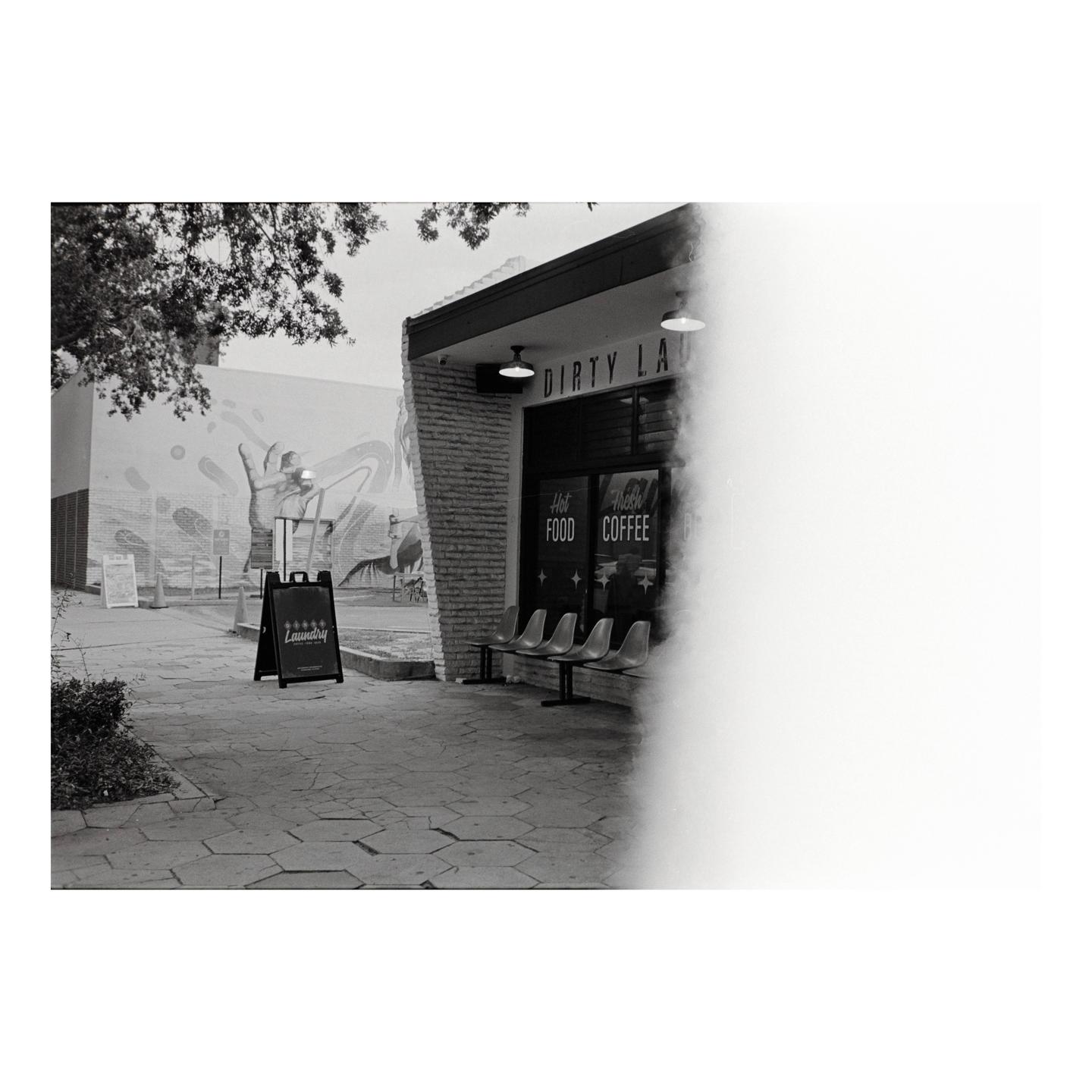 A black and white street view shows a laundry and coffee shop with a mural on the adjacent building.