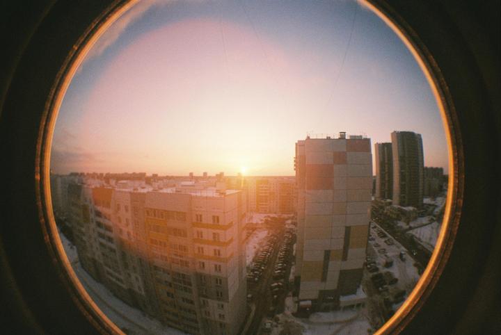 A fisheye view of a city at sunset shows apartment buildings and a parking lot covered in snow.