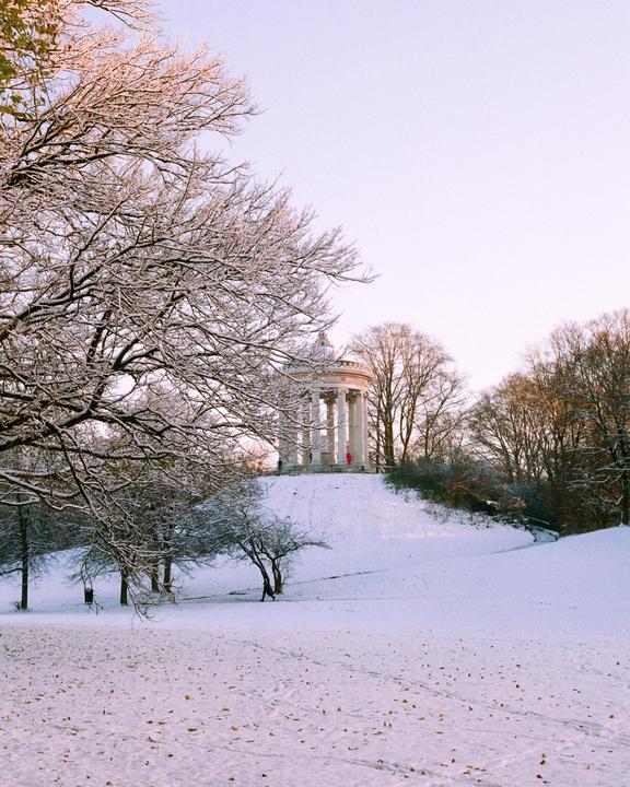 A snow-covered landscape features a classical temple on a hill, with bare trees in the foreground and background.