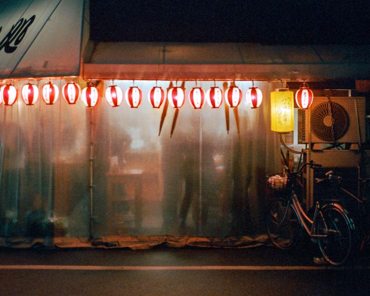 A row of red and white lanterns hangs above a translucent curtain, with a bicycle parked to the right and a yellow lantern hanging nearby.