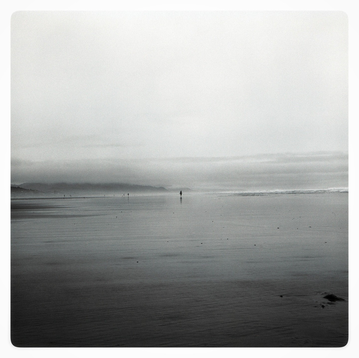 A solitary figure walks on a vast, wet beach under a hazy sky.