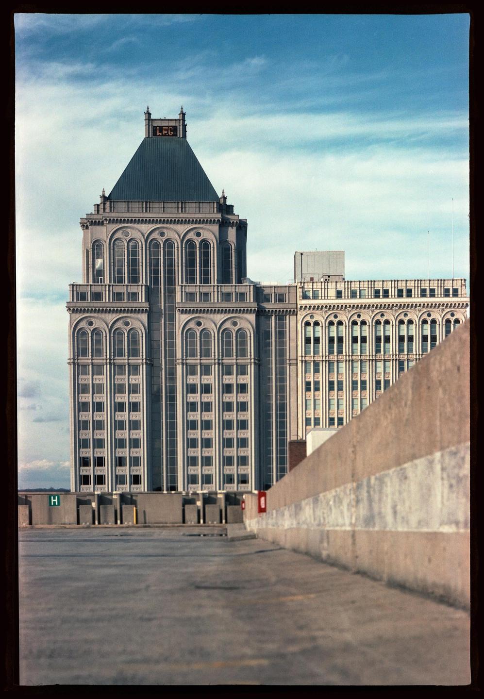 A tall building with a pyramidal roof and many arched windows stands against a blue sky with wispy clouds, viewed from a concrete parking structure.