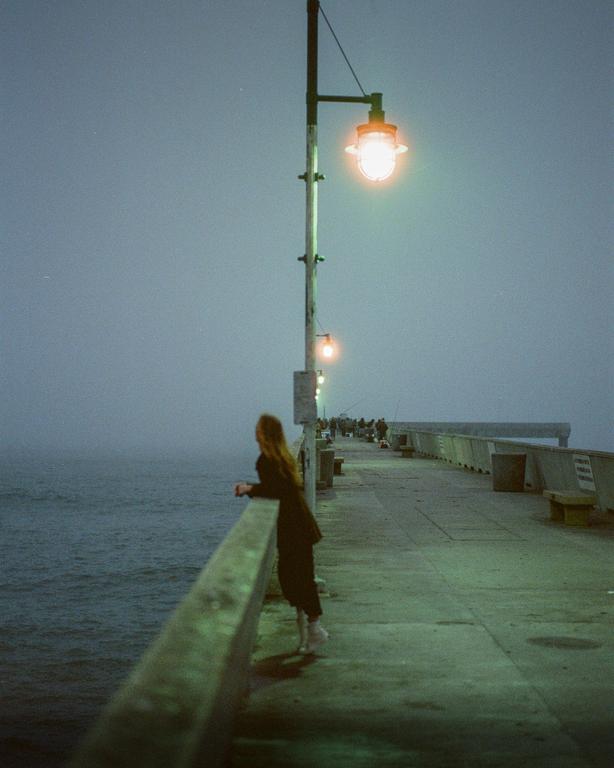 A woman stands on a pier at night, looking out at the water.