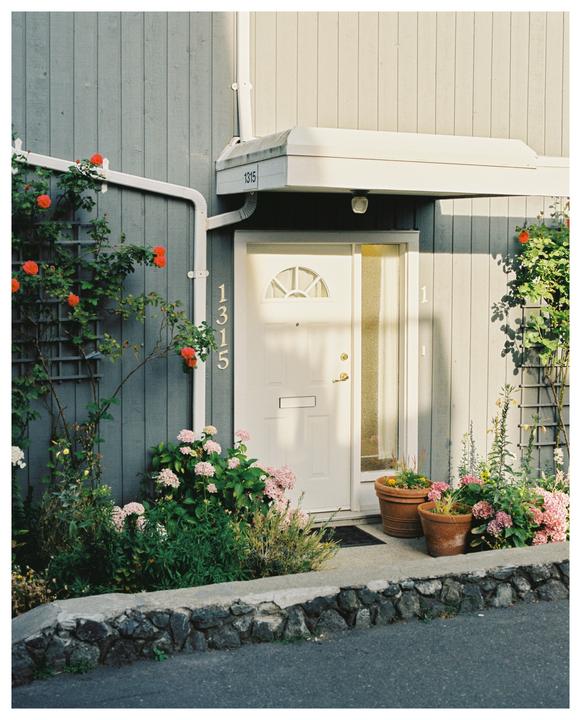 A white door with the number 1315 is surrounded by various plants and flowers next to a gray building.