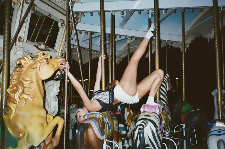 A woman in white shorts and a black top is upside down on a carousel horse at night.