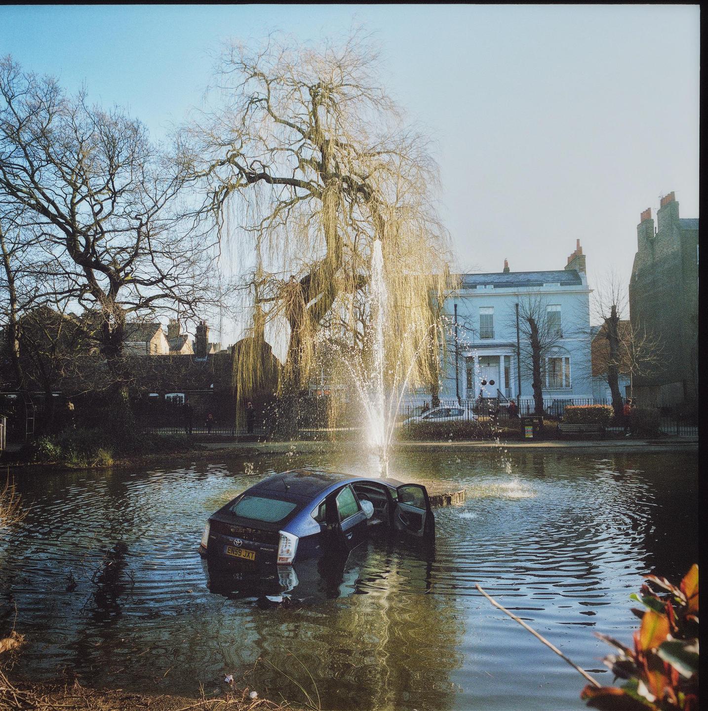 A blue car is submerged in a pond with a fountain spraying water next to it, surrounded by trees and a large house in the background.