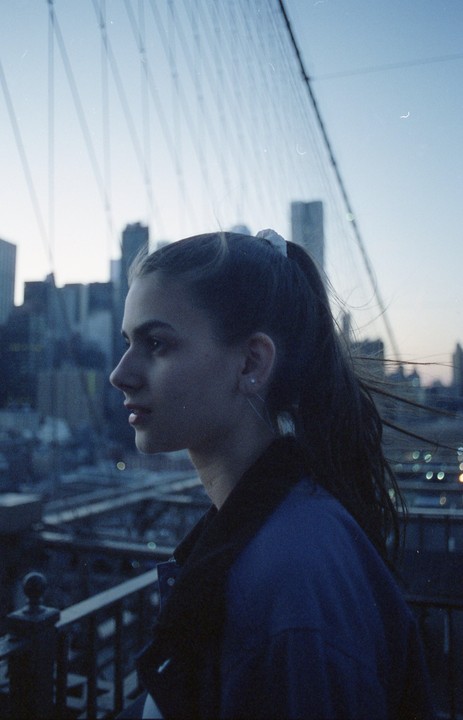 A woman with a ponytail stands on a bridge overlooking a city skyline at dusk.