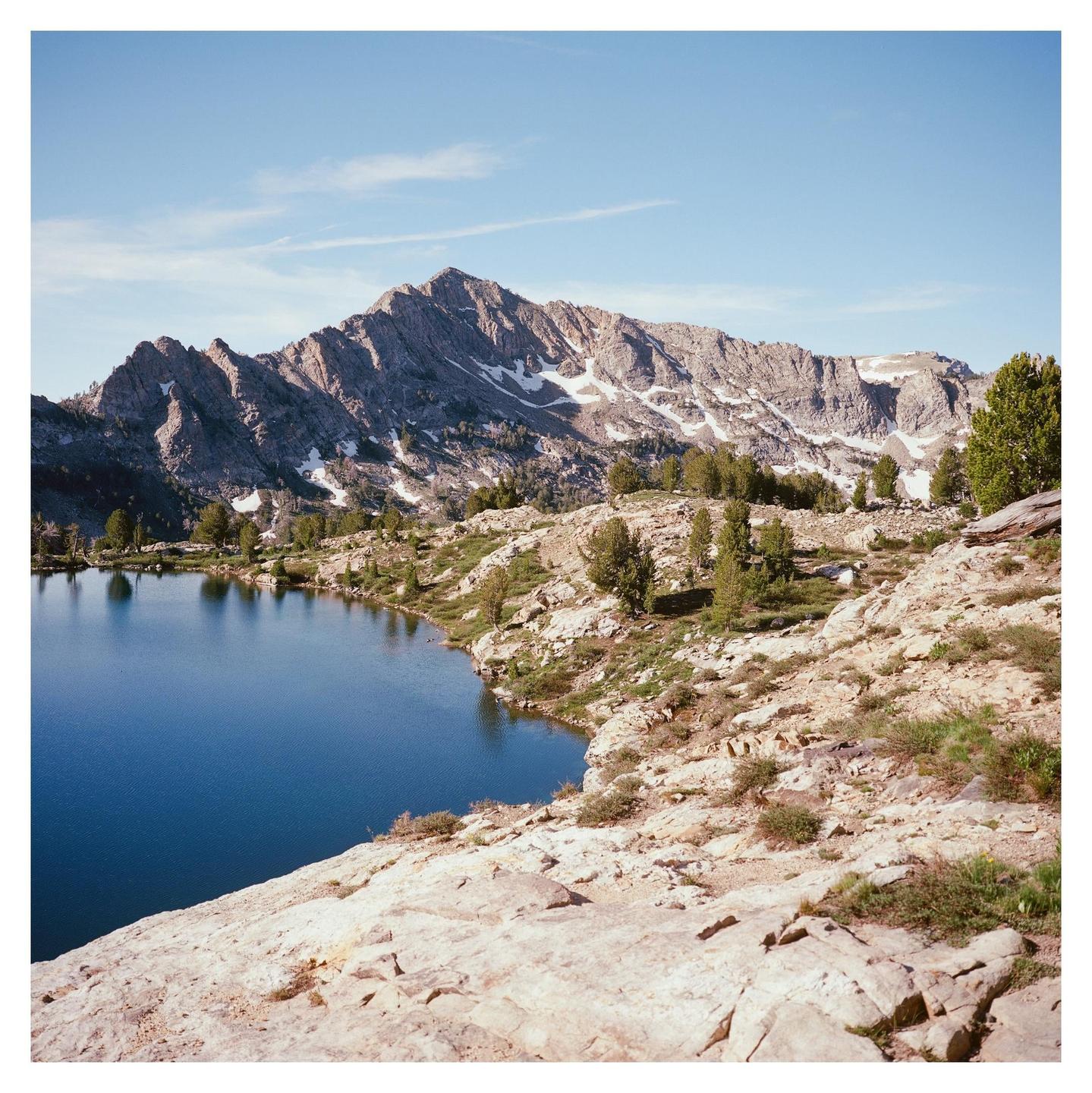 A clear blue lake is nestled among rocky terrain with snow-capped mountains in the background under a bright sky.